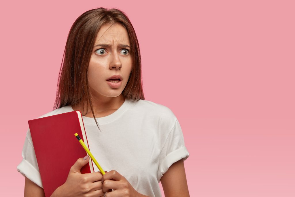 Cómo dejar de procrastinar en una oposición: guía práctica de psicopedagogía del rendimiento 7 surprised horrified young woman with dark straight hair holds red textbook pencil feels puzzled recieving many tasks