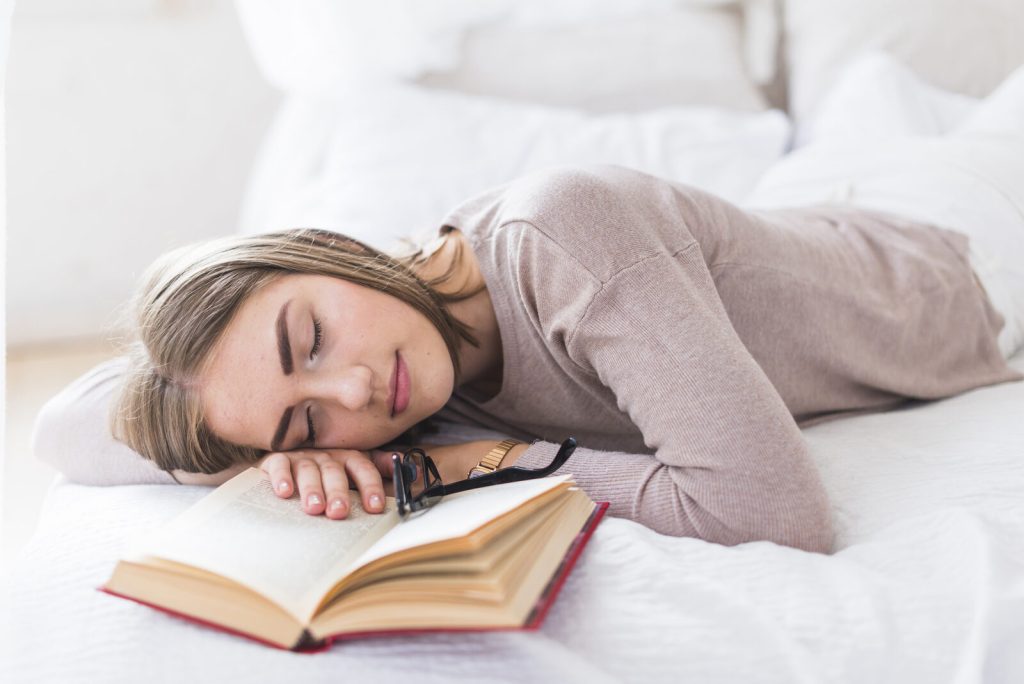 Mujer joven dormida sobre un libro abierto en la cama, símbolo de que dormir consolida lo estudiado